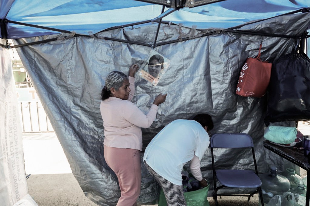 Dos mujeres trabajando en un puesto de mercado, una de pie colgando un cartel, y otra agachada revisando una bolsa.