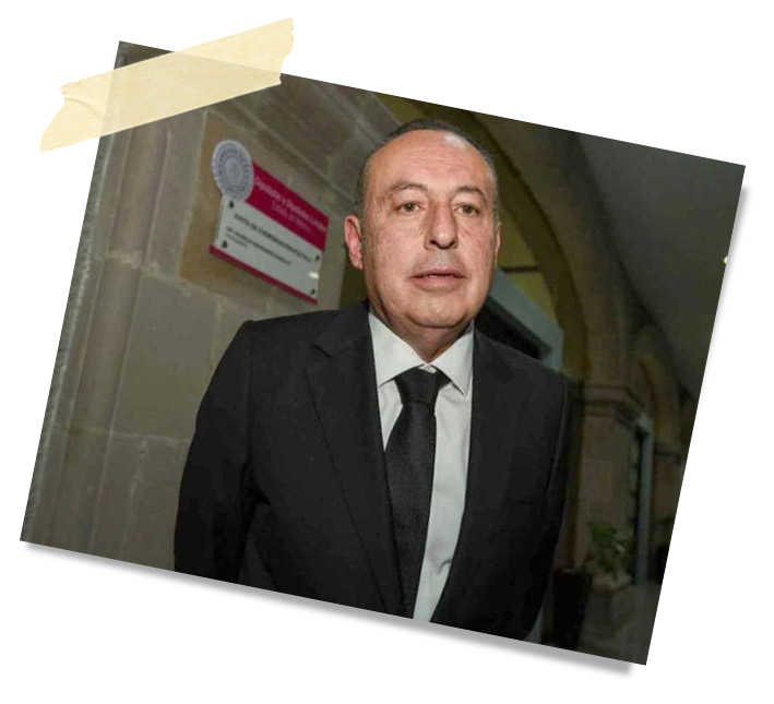 Hombre de pie en un entorno elegante, vistiendo un traje negro y corbata, con una expresión seria. Hay un letrero visible en la pared detrás de él.