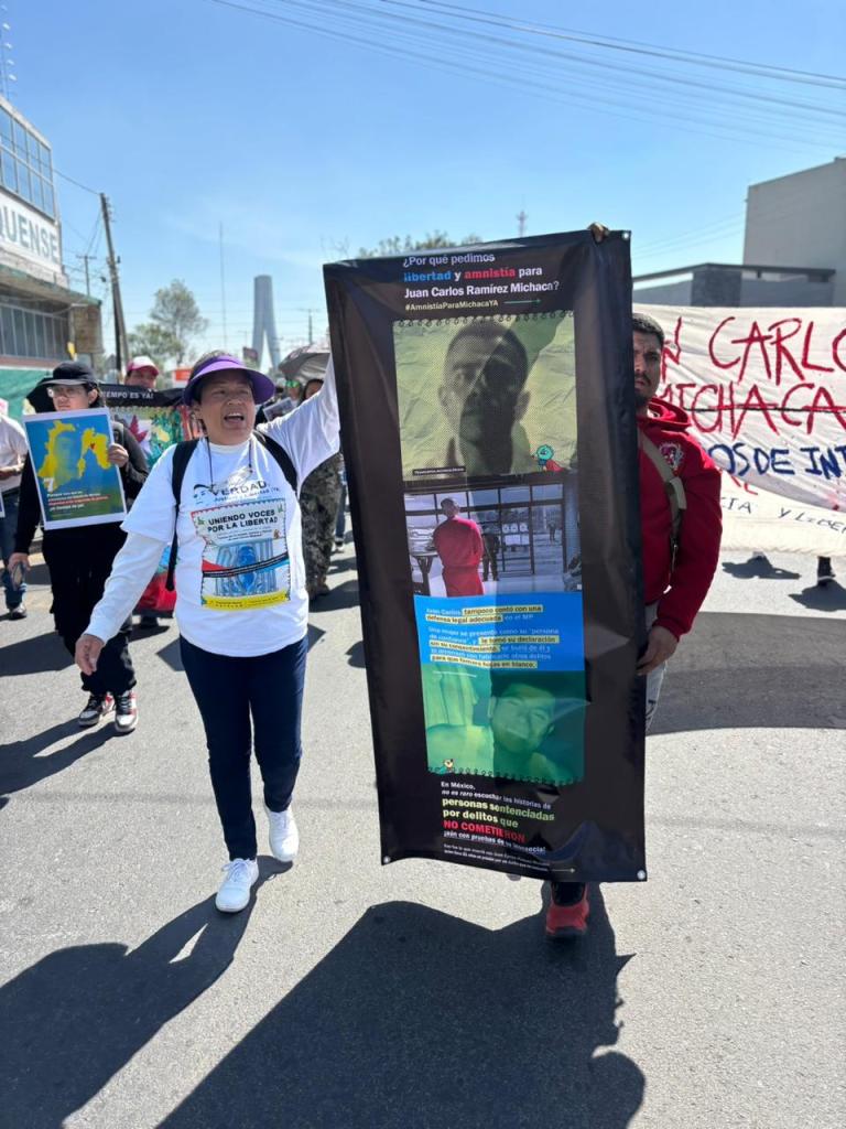 A group of protesters marching in the street, one woman holding a banner calling for freedom and amnesty for Juan Carlos Ramirez Michacan. The banner features images and text related to the cause.