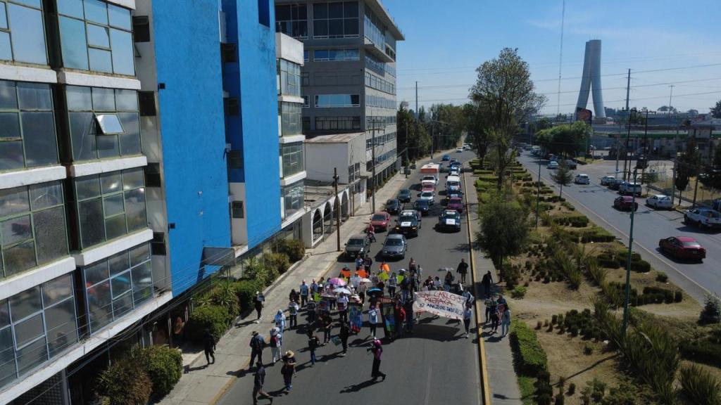 Aerial view of a street in an urban area where a group of people is participating in a protest or march, holding banners and raising signs. Buildings line the street, and cars are parked along the road.