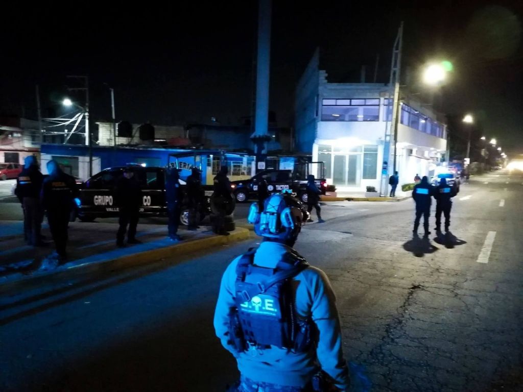 A group of police officers in tactical gear gathering at night, with police vehicles parked nearby on a city street.
