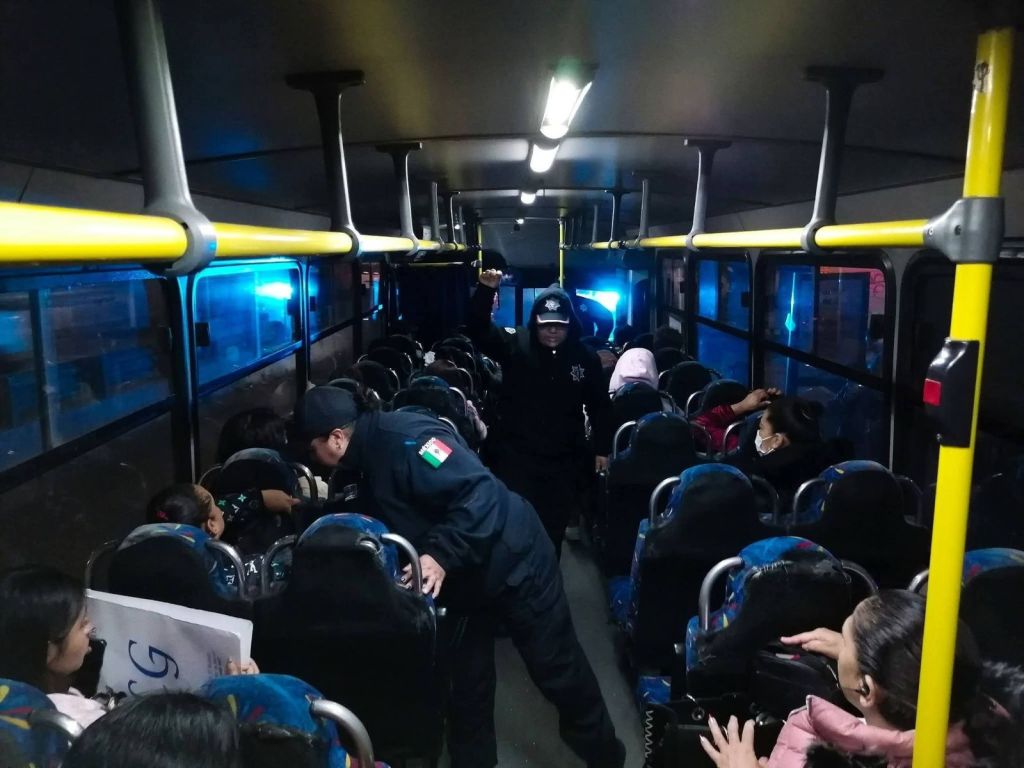 Interior of a bus with several passengers seated, including a police officer standing in the aisle and another officer interacting with passengers, under dim lights with blue illumination.