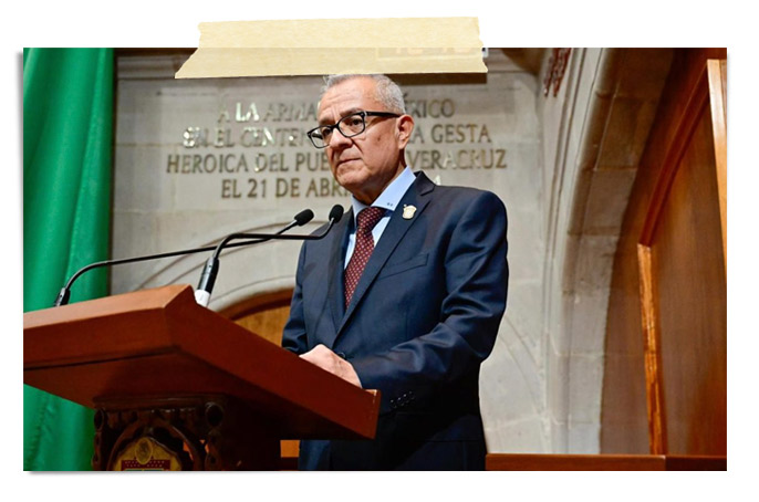 Un hombre en traje y gafas de lectura, de pie en un podio, mientras habla. Al fondo, una pared con un mural y una bandera. La escena parece ser un evento formal o una ceremonia.