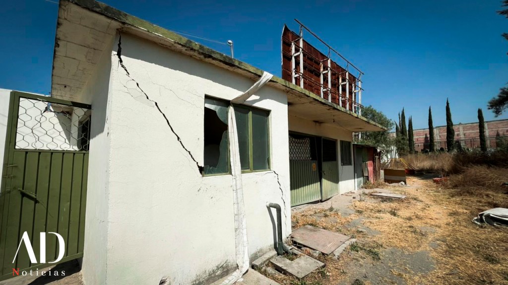 Vista de un edificio deteriorado con ventanas quebradas, puerta verde y vegetación seca alrededor.