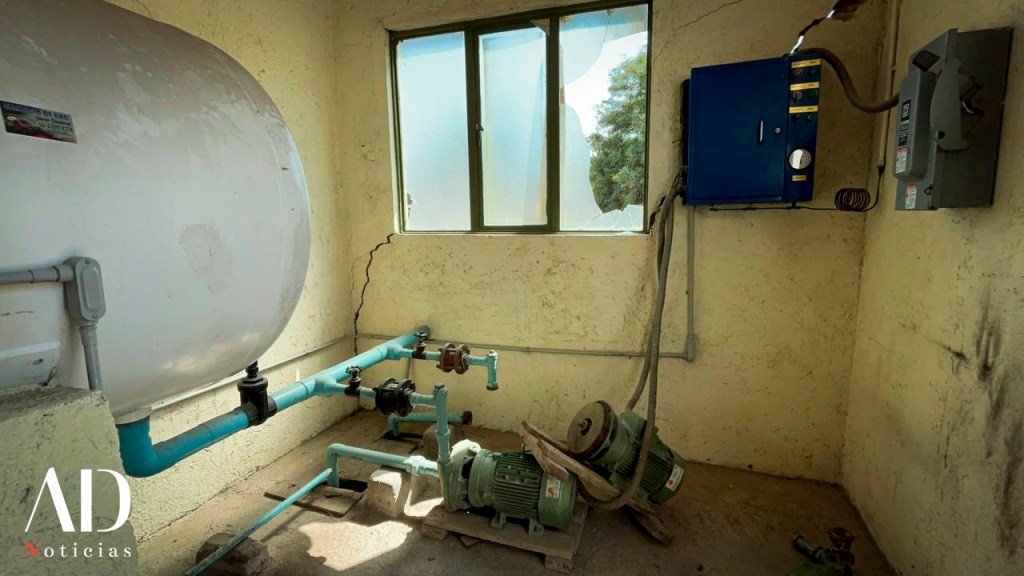 Interior de un cuarto técnico con un tanque blanco de agua, tuberías azules, dos motores eléctricos en el suelo y un panel de control azul en la pared.