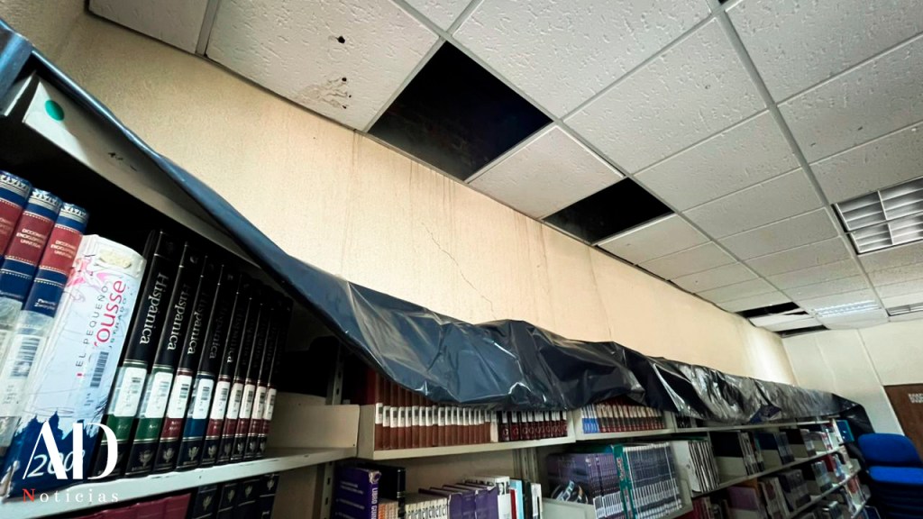 Estantería de libros en una biblioteca con un plástico negro cubriendo la parte superior, y daños visibles en el techo, incluyendo paneles faltantes.