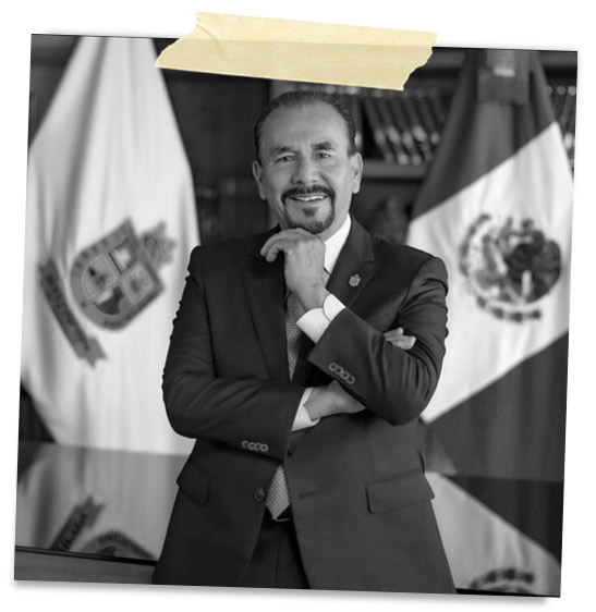 Retrato en blanco y negro de un hombre sonriente con traje, de pie con los brazos cruzados, frente a banderas de México y un fondo de estanterías.
