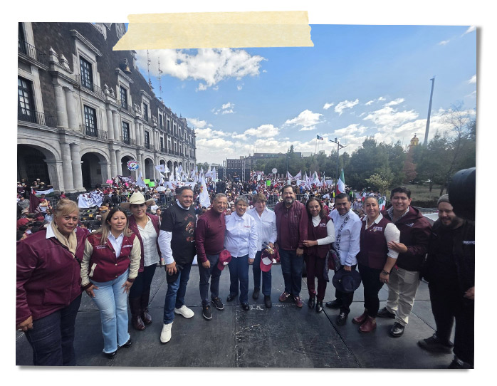 Un grupo de personas posando para una foto en una manifestación al aire libre, con banderas y pancartas visibles en el fondo. El lugar es un espacio público, y se observa un edificio histórico detrás de ellos.