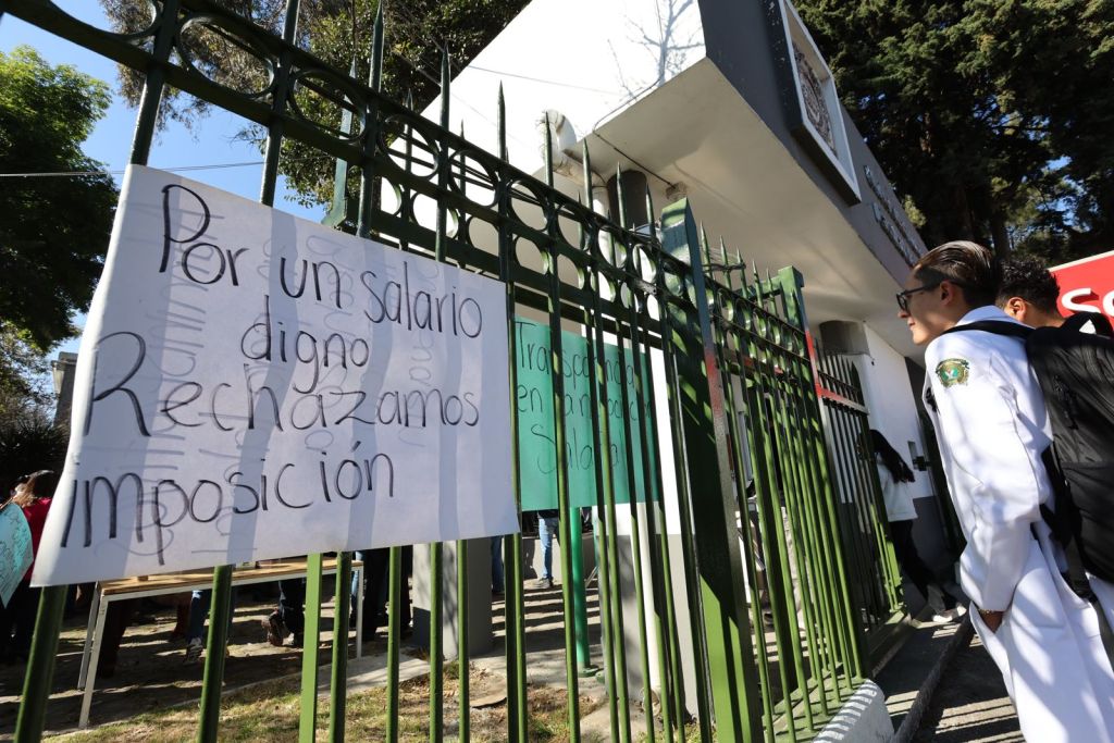 A protest scene outside a building, featuring a sign that reads 'Por un salario digno Rechazamos imposición' attached to a green fence, with a person in a white uniform looking at the sign.