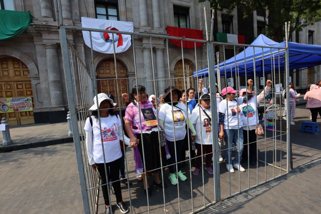 Group of women holding hands inside a metal cage during a protest, wearing white T-shirts with images and messages, with a backdrop of a building and banners.