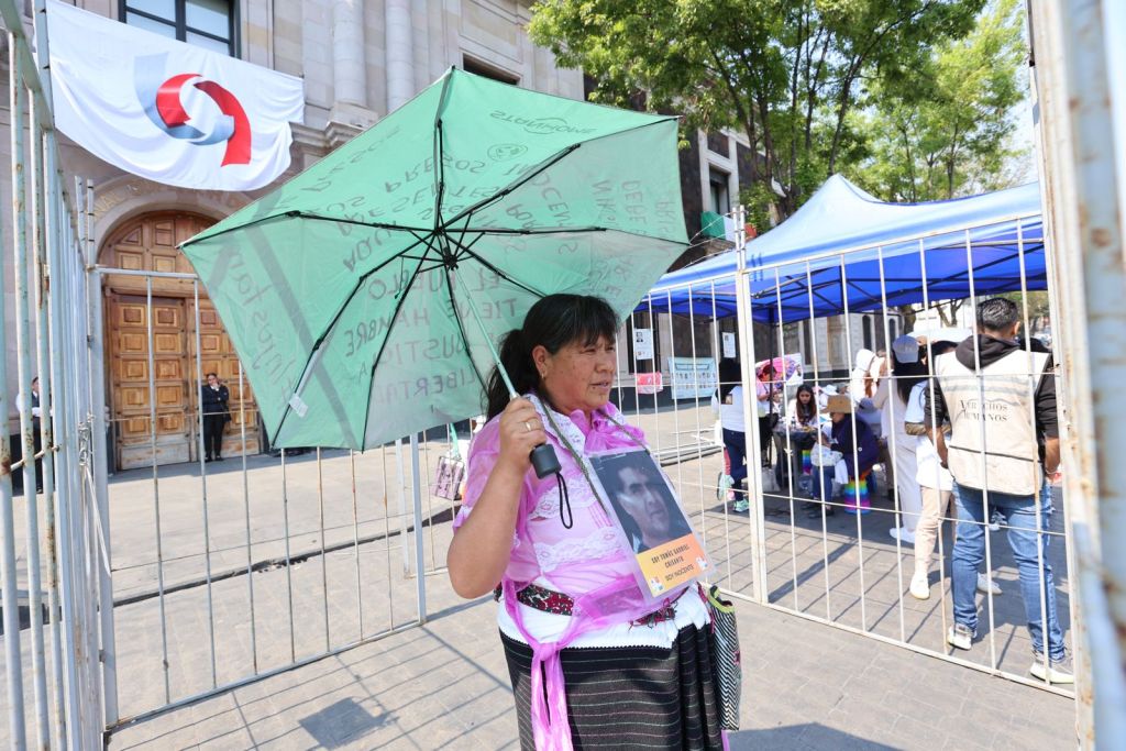 A woman holding a green umbrella stands outside a gated area, wearing traditional clothing with a photo hanging around her neck. In the background, people gather under blue tents near a large building.