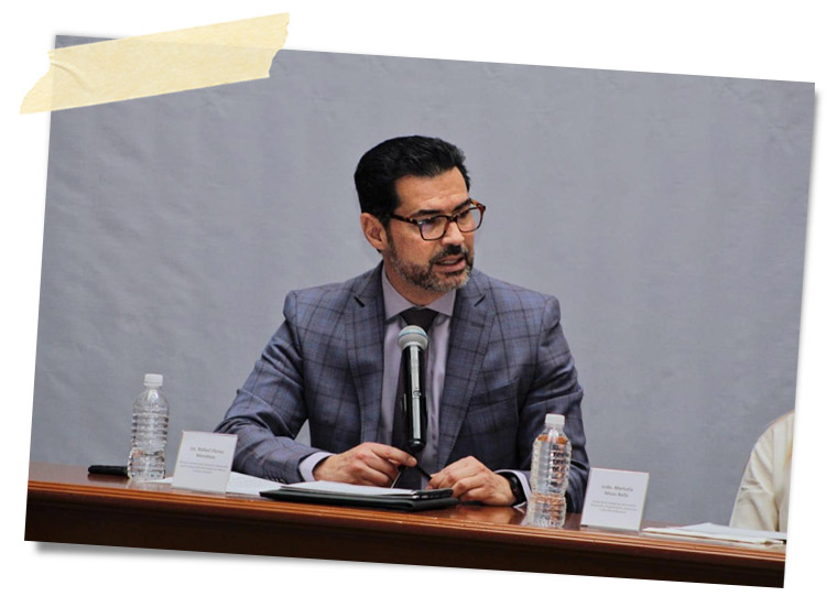 Hombre con gafas sentado en una mesa con micrófono, hablando durante un evento, con botellas de agua y documentos visibles.