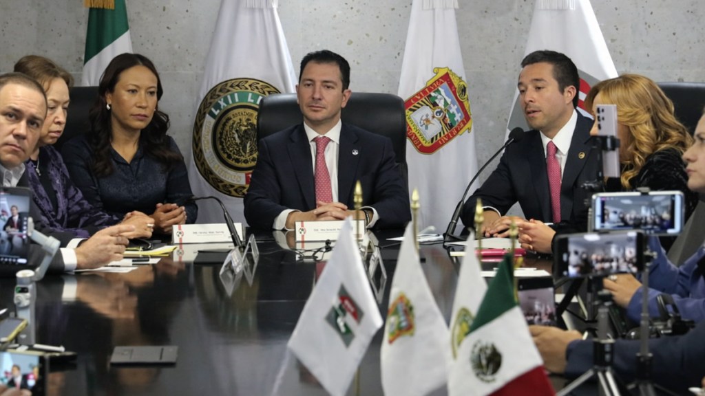 Reunión de funcionarios en una mesa, con banderas de México y otros estados al fondo, discutiendo temas importantes.
