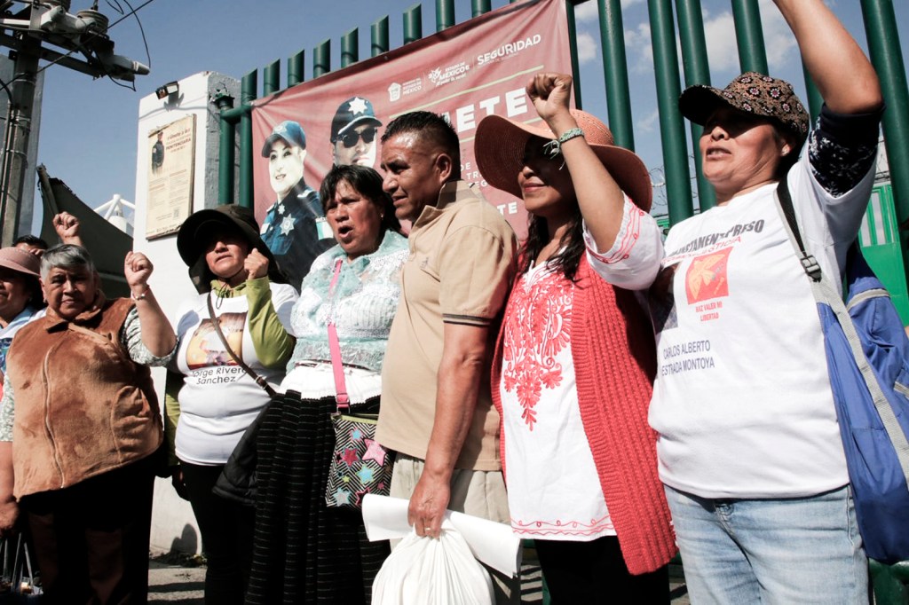Group of individuals raising their fists in solidarity during a demonstration, with various expressions of determination, in front of a green fence and a poster featuring law enforcement.