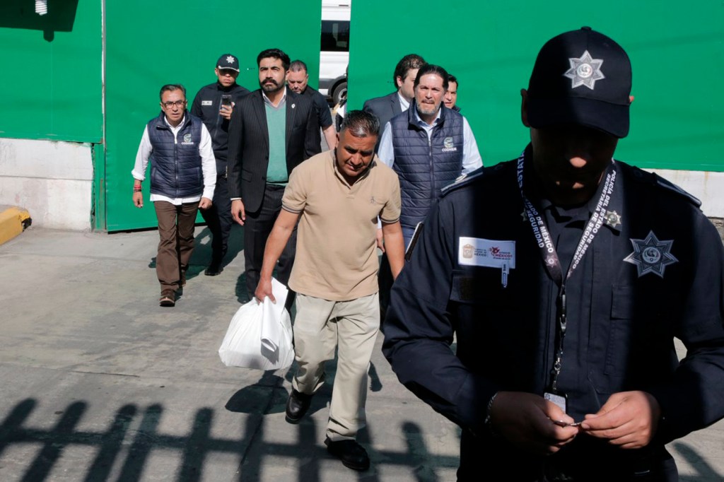 A group of individuals, including a police officer in uniform, walking through a gate, with one person carrying a bag. The background features a green wall and various people in formal attire.