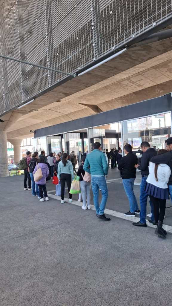 A long line of people waiting outside a building, standing on a concrete pathway under a modern architectural structure.
