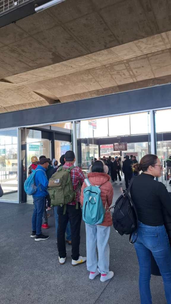 A line of people waiting outside a building, with a mix of casual attire and backpacks, under a concrete overhang.