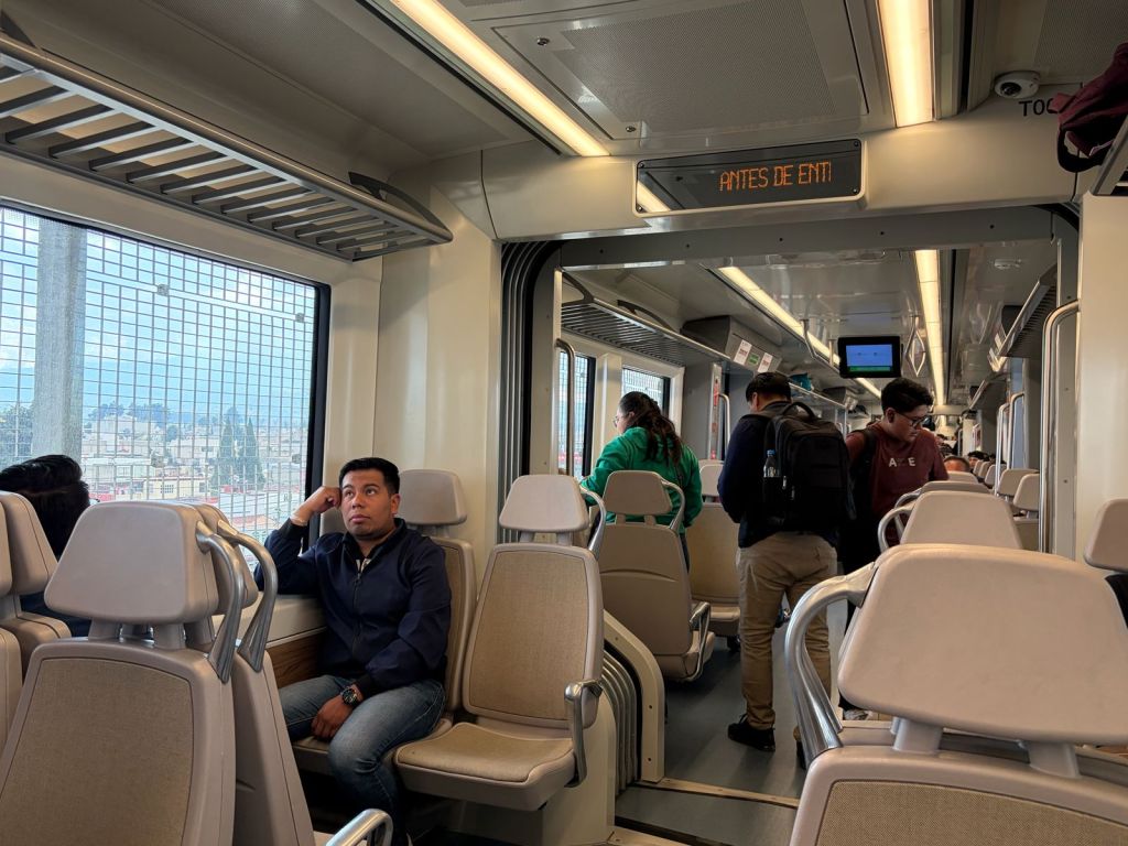 Interior of a public transportation train with passengers seated and standing, featuring a man looking out the window, and a digital display showing 'ANTES DE ENTI'.