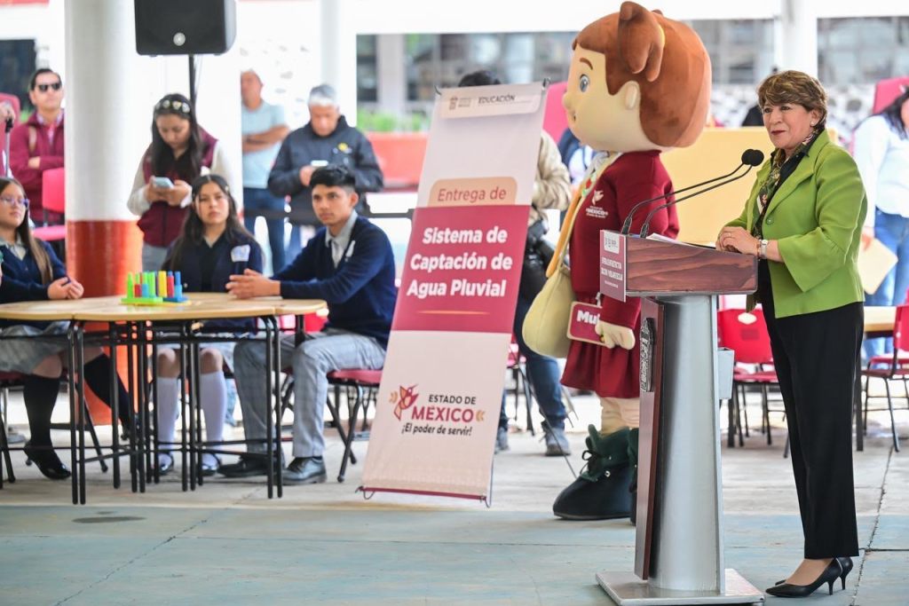 Evento de entrega del sistema de captación de agua pluvial, con una mujer hablando en un podio y un personaje de mascota al lado, mientras estudiantes observan desde sus asientos.