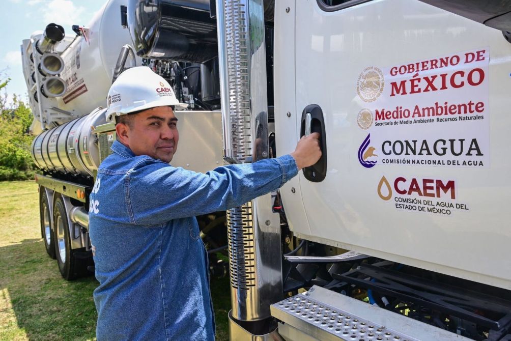 A worker in a blue shirt and white hard hat is standing next to a large truck with Spanish logos related to the Mexican government and environmental services.