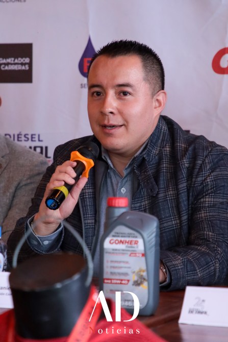 A man speaking into a microphone at a press conference, with promotional materials and a bottle of motor oil on the table.