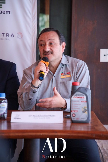 A man speaking into a microphone at a conference table, wearing a gray shirt with an orange logo. A bottle of oil is visible on the table next to him.
