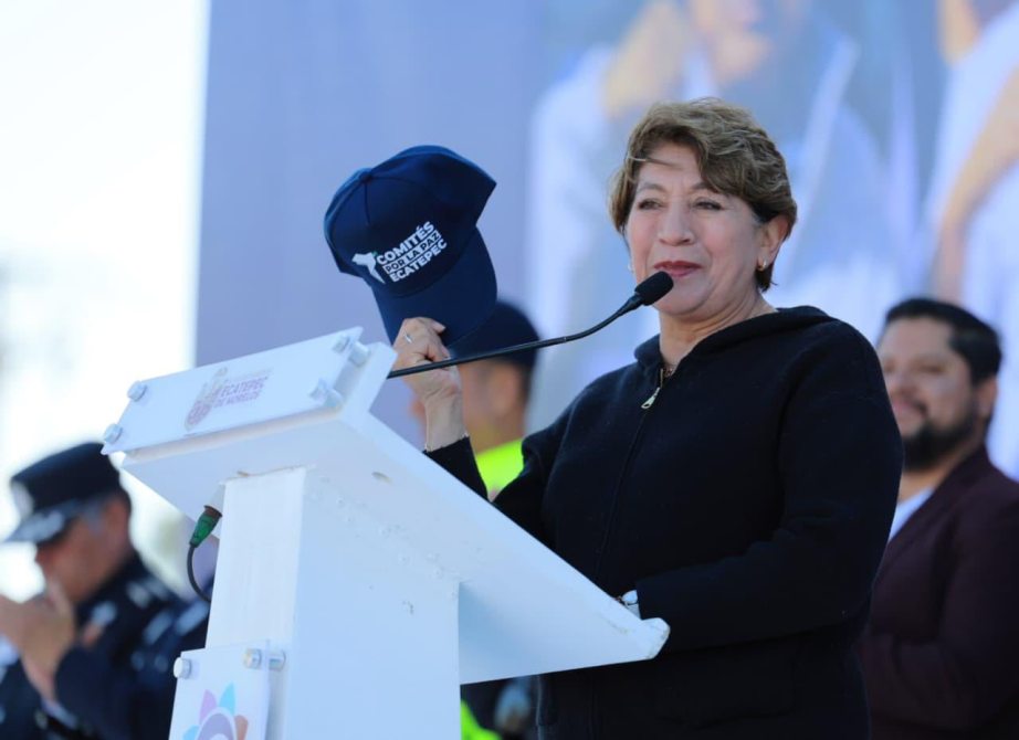 A woman speaking at a podium, holding a blue hat with the text 'COMITES DE SEGURIDAD.' She appears to be addressing an audience, with police officers visible in the background.