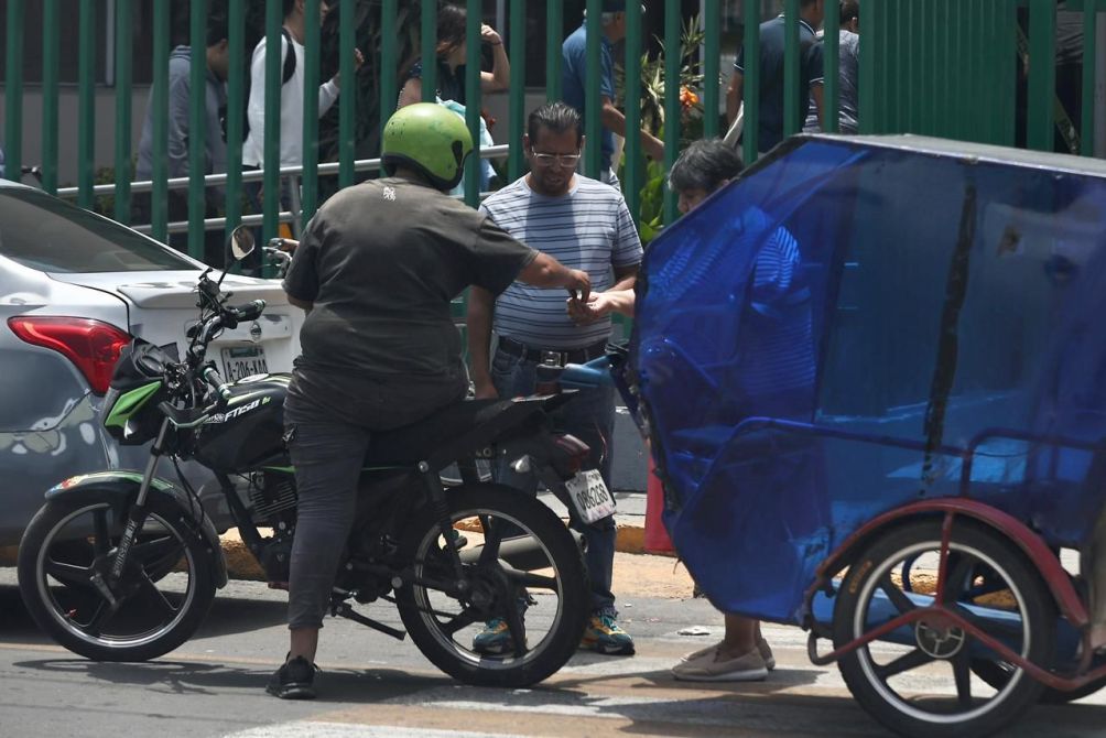 A motorcycle rider in a helmet interacts with a person in a striped shirt on a busy street, while a tricycle is partially visible in the foreground.