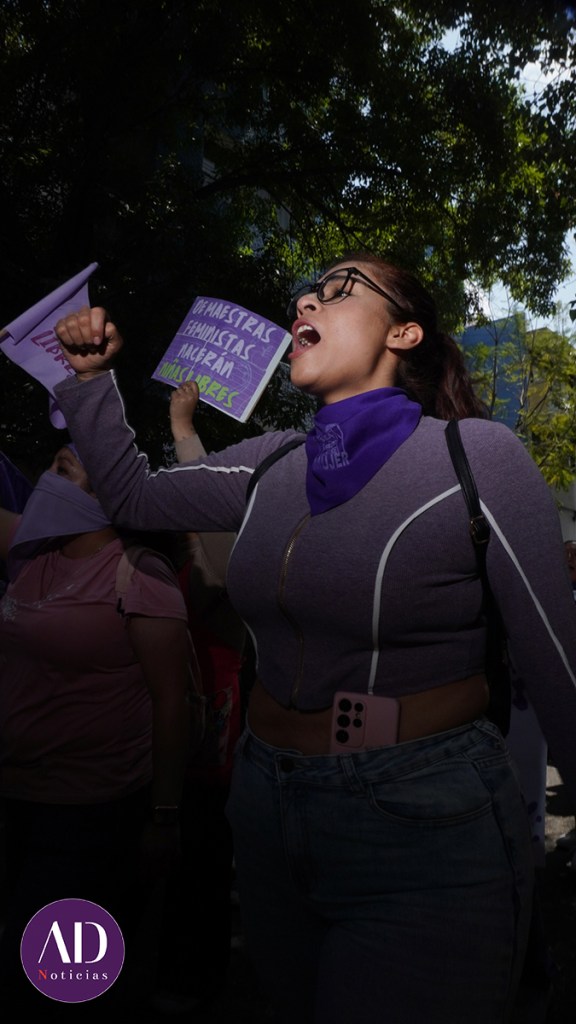 Mujer protestando con el puño en alto, usando un pañuelo morado y gritando con pasión, mientras sostiene un cartel en un ambiente de manifestación.