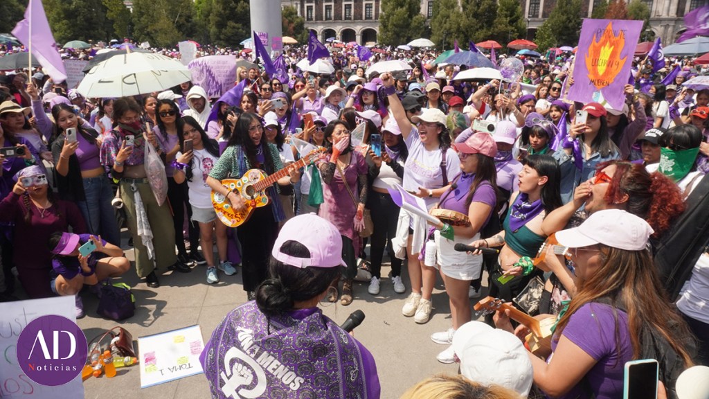Mujeres participando en una manifestación, vistiendo ropa morada y rosa, algunos sosteniendo pancartas y otros tocando instrumentos musicales. Se observa una gran multitud detrás, con paraguas y banderas moradas.
