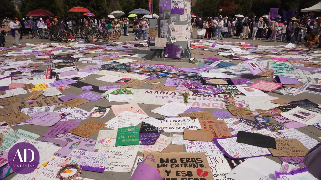 Vista de una manifestación con carteles sobre el suelo, muchos de ellos en tonos morados, en un espacio público, donde se observa a personas reunidas y sosteniendo paraguas.