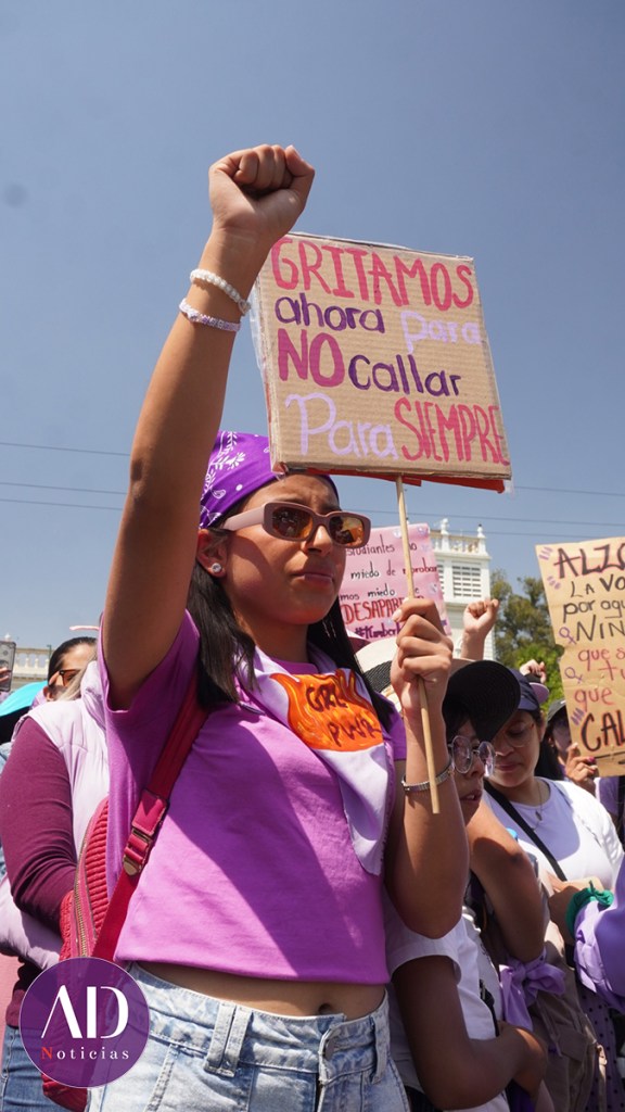 Joven levantando el puño en una manifestación, sosteniendo un cartel que dice 'GRITAMOS ahora para NO callar Para SIEMPRE'.