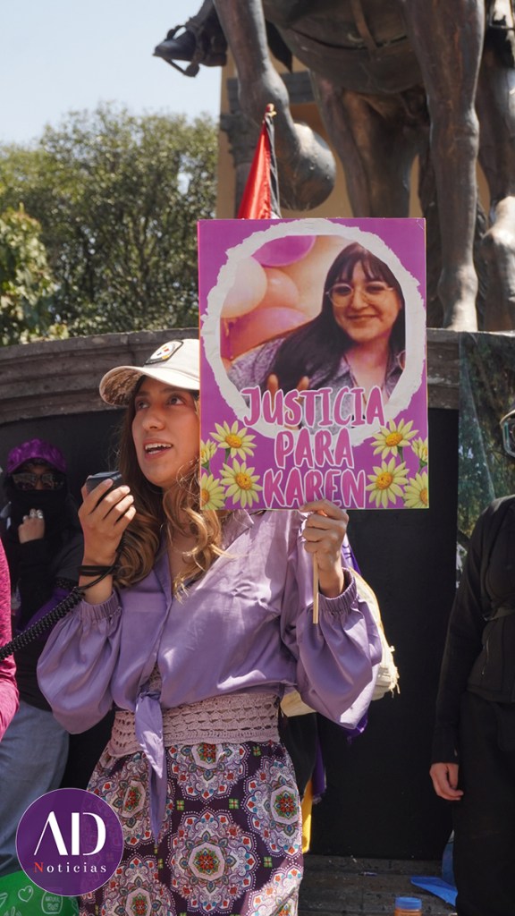 Una joven sostiene un cartel que dice 'Justicia para Karen' en una manifestación, con una imagen de Karen en el fondo.