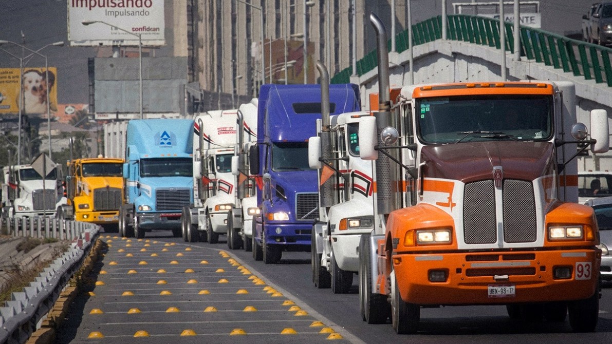 A line of colorful freight trucks traveling on a busy road, with buildings and advertisements in the background.