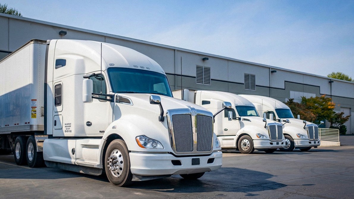 Three white semi-trucks parked in front of a warehouse on a sunny day.