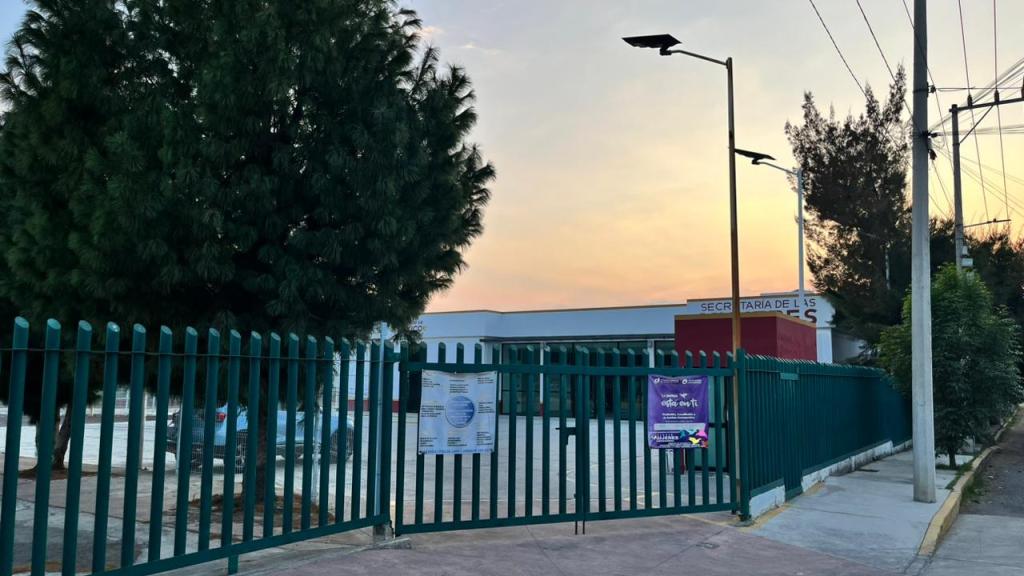 Fachada de una escuela rodeada por una cerca verde, con árboles y un atardecer en el cielo.
