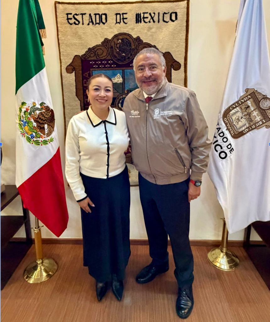 Un hombre y una mujer posan sonrientes en un edificio gubernamental de México, con banderas de México y del Estado de México de fondo.