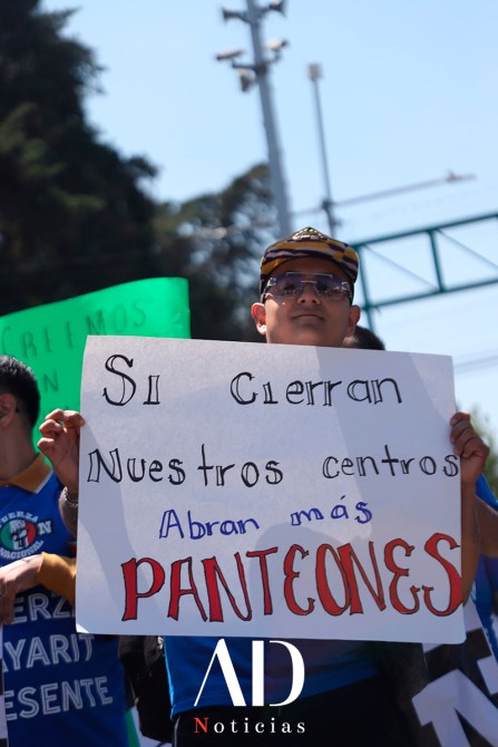 A participant in a protest holds a sign that reads 'Si cierran nuestros centros, abran más panteones' while wearing sunglasses and a cap.