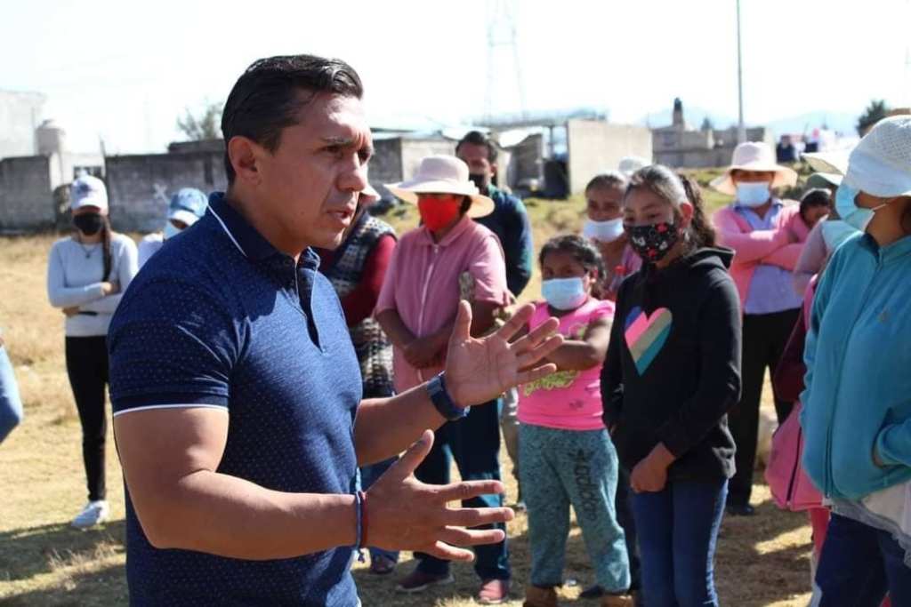 Hombre hablando enérgicamente frente a un grupo de personas, muchas de las cuales están escuchando atentamente en un campo al aire libre.