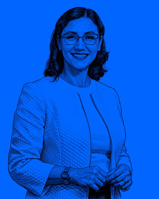 Retrato de una mujer con gafas, sonriendo y usando una chaqueta blanca, sobre un fondo azul.