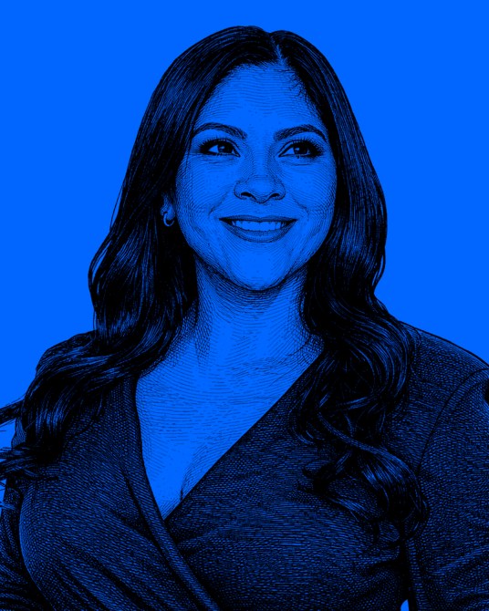 Retrato de una mujer con cabello largo y oscuro, sonriendo frente a un fondo azul. Ella lleva una blusa de color oscuro.