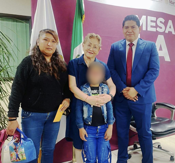 Cuatro personas posando para la foto en un evento, con banderas y un fondo que muestra el texto 'MESA'. Dos mujeres, un niño y un hombre, todos sonriendo.