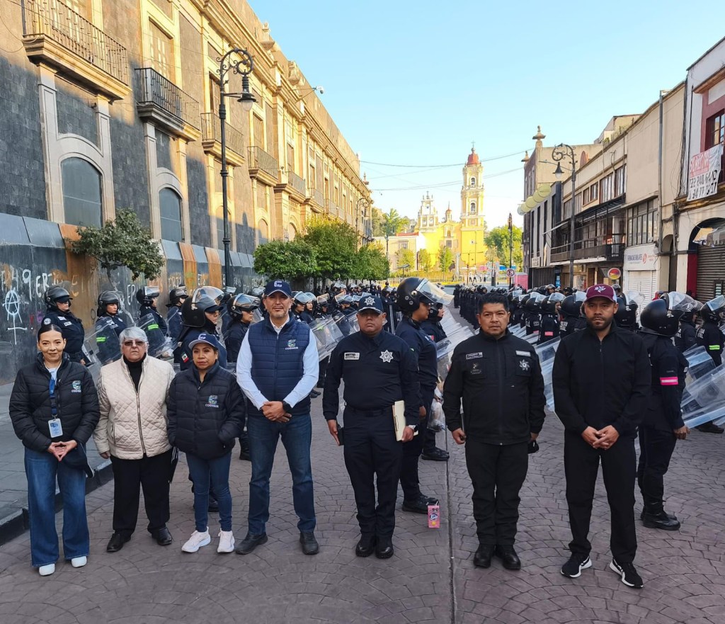Grupo de personas frente a un cordón de policías en una calle, con edificios históricos de fondo, durante un despliegue de seguridad.