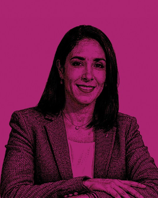 Retrato de una mujer sonriendo, vestida con un blazer, contra un fondo de color magenta.