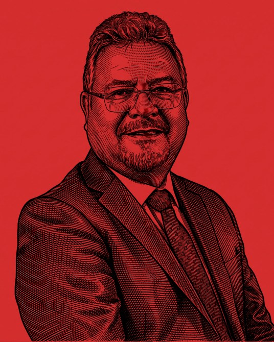 Retrato de un hombre de negocios con cabello encanecido, usando traje y gafas, sobre un fondo rojo.