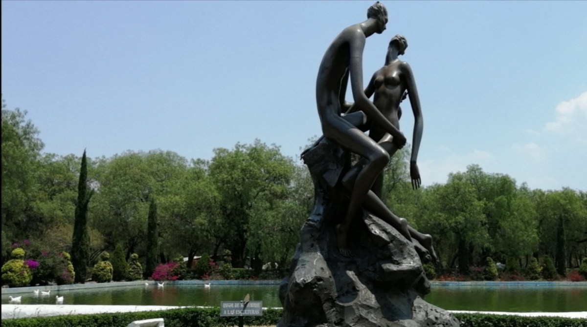 Escultura de dos figuras humanas, una masculina y una femenina, sentadas sobre una roca en un entorno natural con árboles y un estanque al fondo.