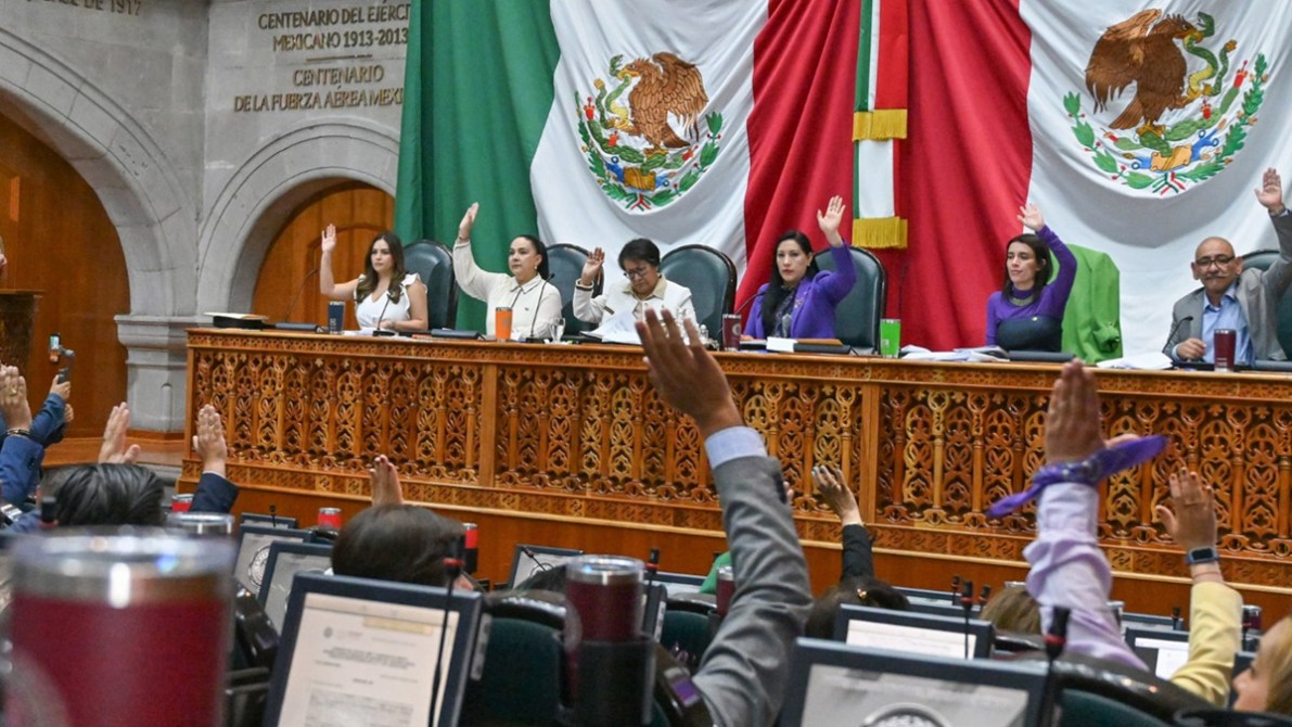 A parliamentary session in progress, with multiple lawmakers raising their hands in a vote. The backdrop features the Mexican flag and a decorative wooden panel.
