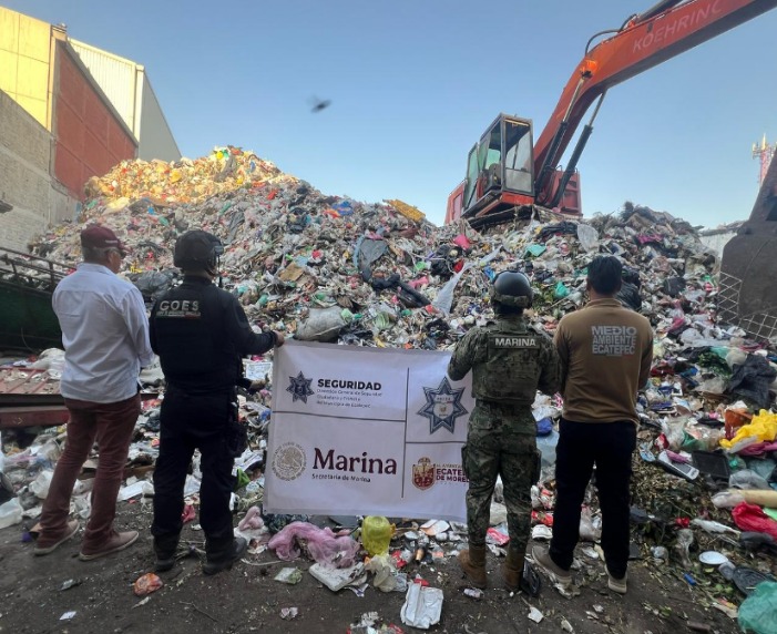 A group of officials standing in front of a large pile of trash, observing a crane in operation at a waste management site.