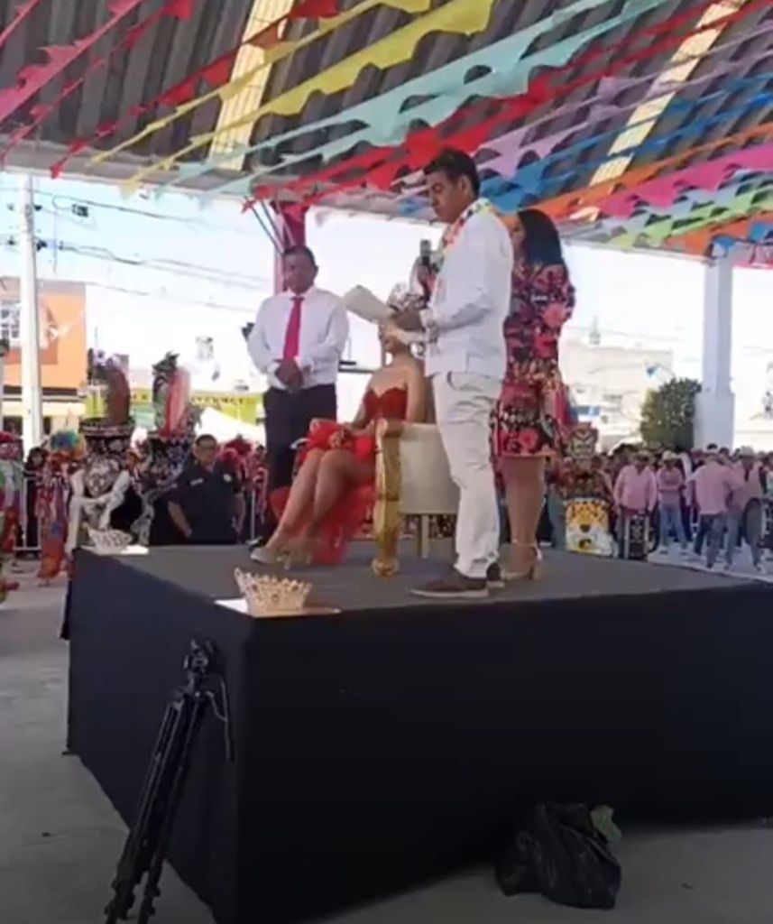 Evento en un escenario con decoraciones coloridas, donde un hombre está entregando un discurso frente a una mujer sentada en una silla, mientras otras personas lo observan.