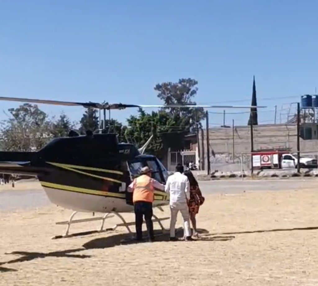 Un grupo de personas se acerca a un helicóptero negro con detalles amarillos en un área al aire libre, con cielo despejado y árboles al fondo.